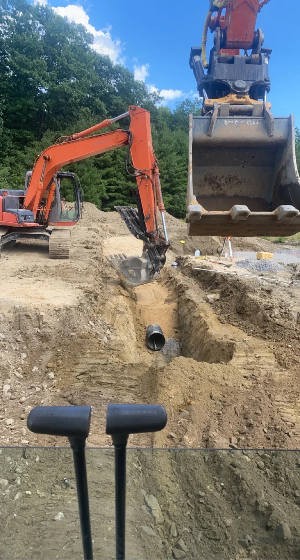 Excavator placing drainage pipe in trench with dump truck and materials on site