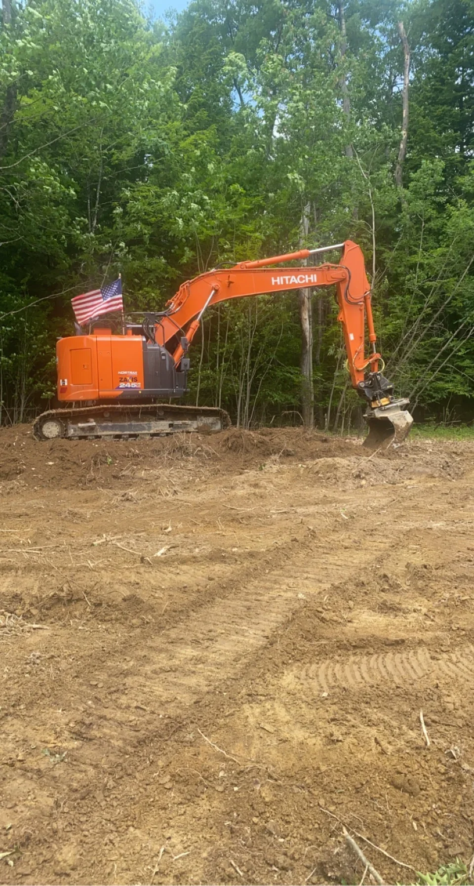 Orange Hitachi excavator with bucket on tracked chassis grading cleared dirt lot surrounded by trees