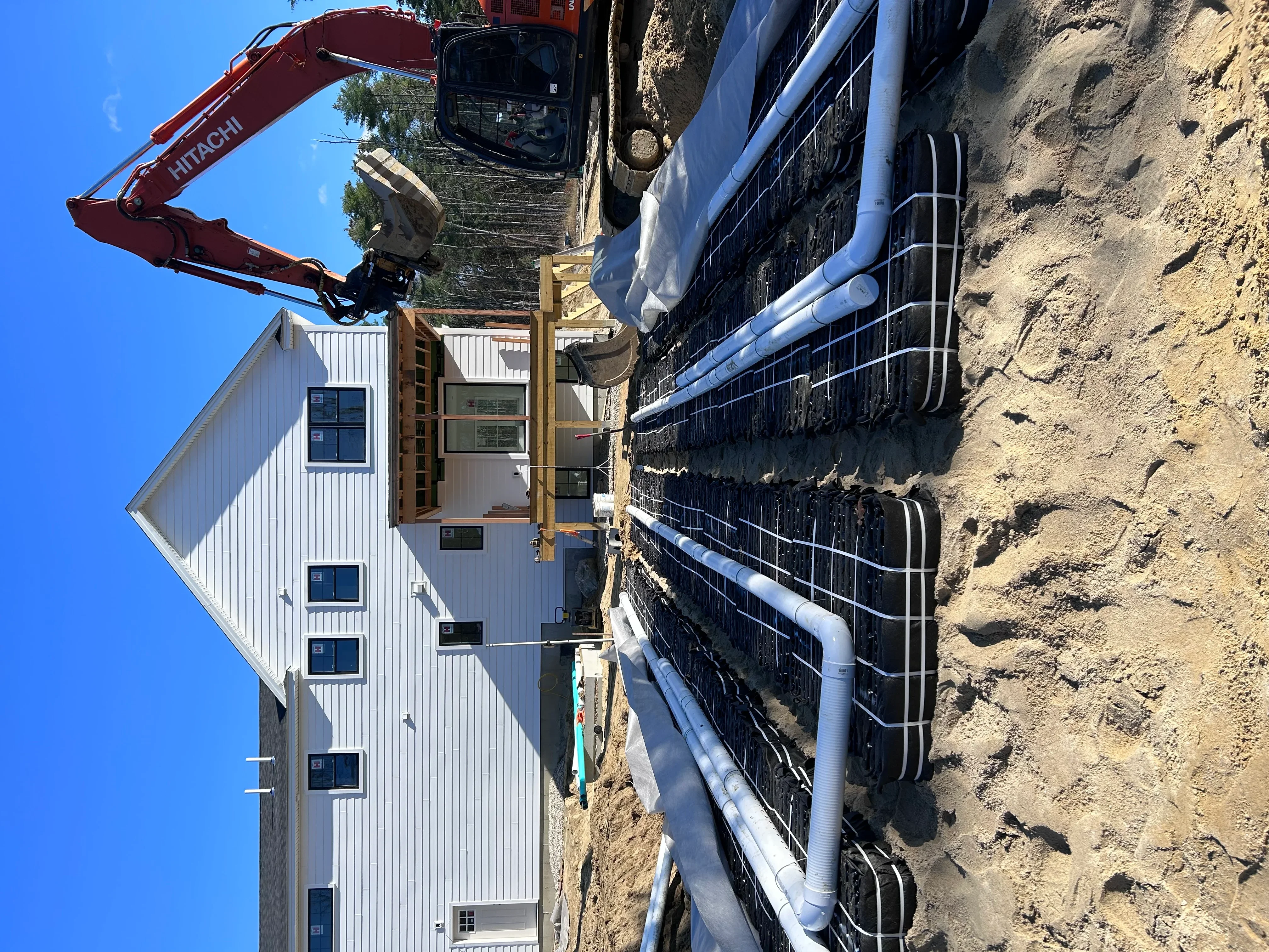 Hitachi excavator grading dirt site next to new metal-sided commercial building with blue drainage pipes