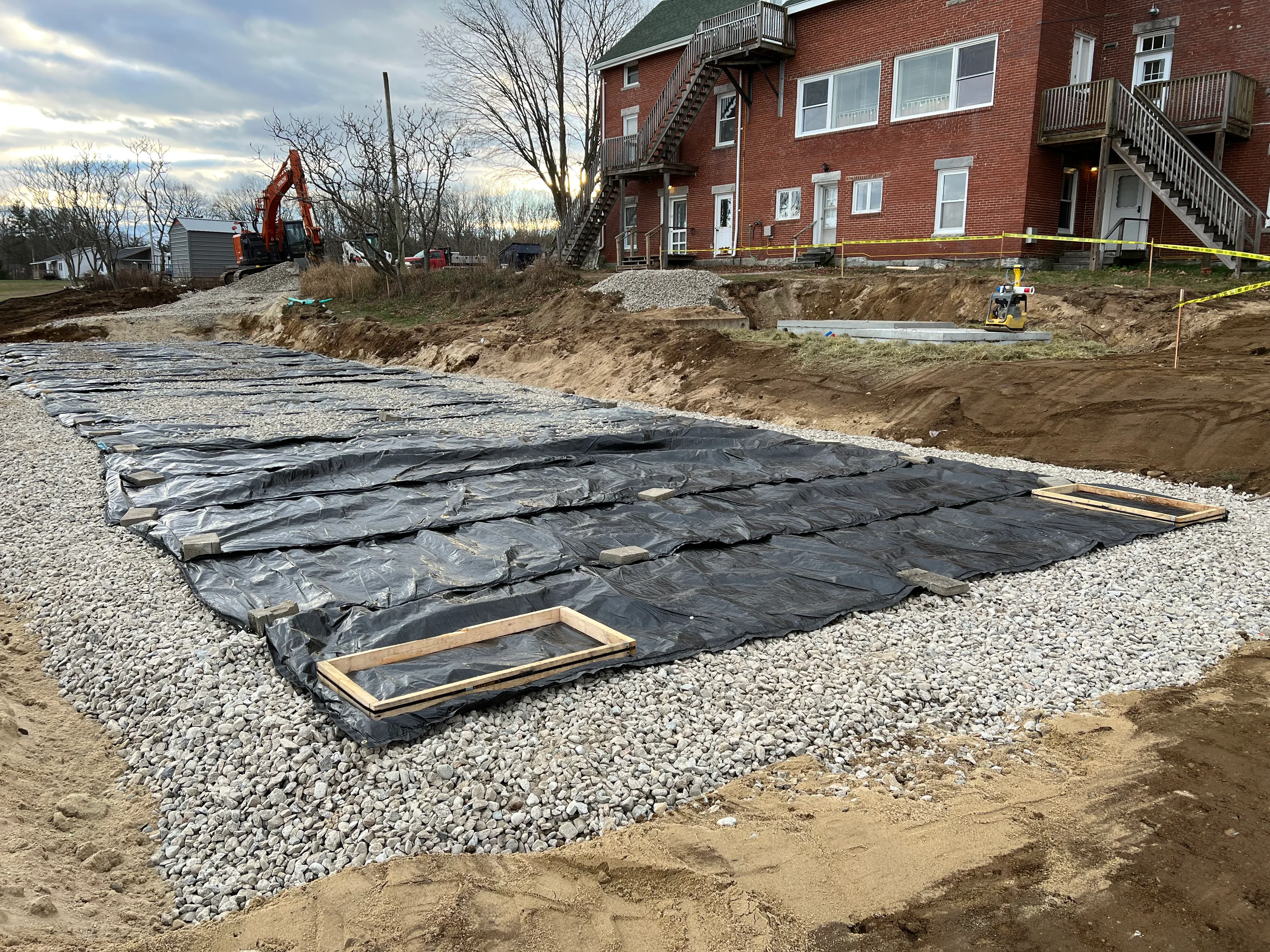 Excavated foundation pit with geotextile membrane, gravel base, and wooden frames near brick building